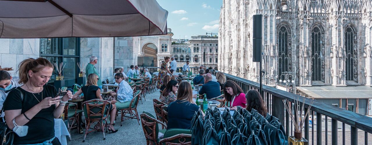 Evento di presentazione della fragranza Duomo Milano di Euthalia Fragrances presso Terrazza Giacomo Arengario, con vista panoramica sul Duomo di Milano. Invitati che partecipano all'evento, seduti ai tavoli all'aperto, con borse regalo su un tavolo in primo piano. Atmosfera elegante e rilassata, perfetta per scoprire la nuova fragranza ispirata alla maestosità del Duomo.