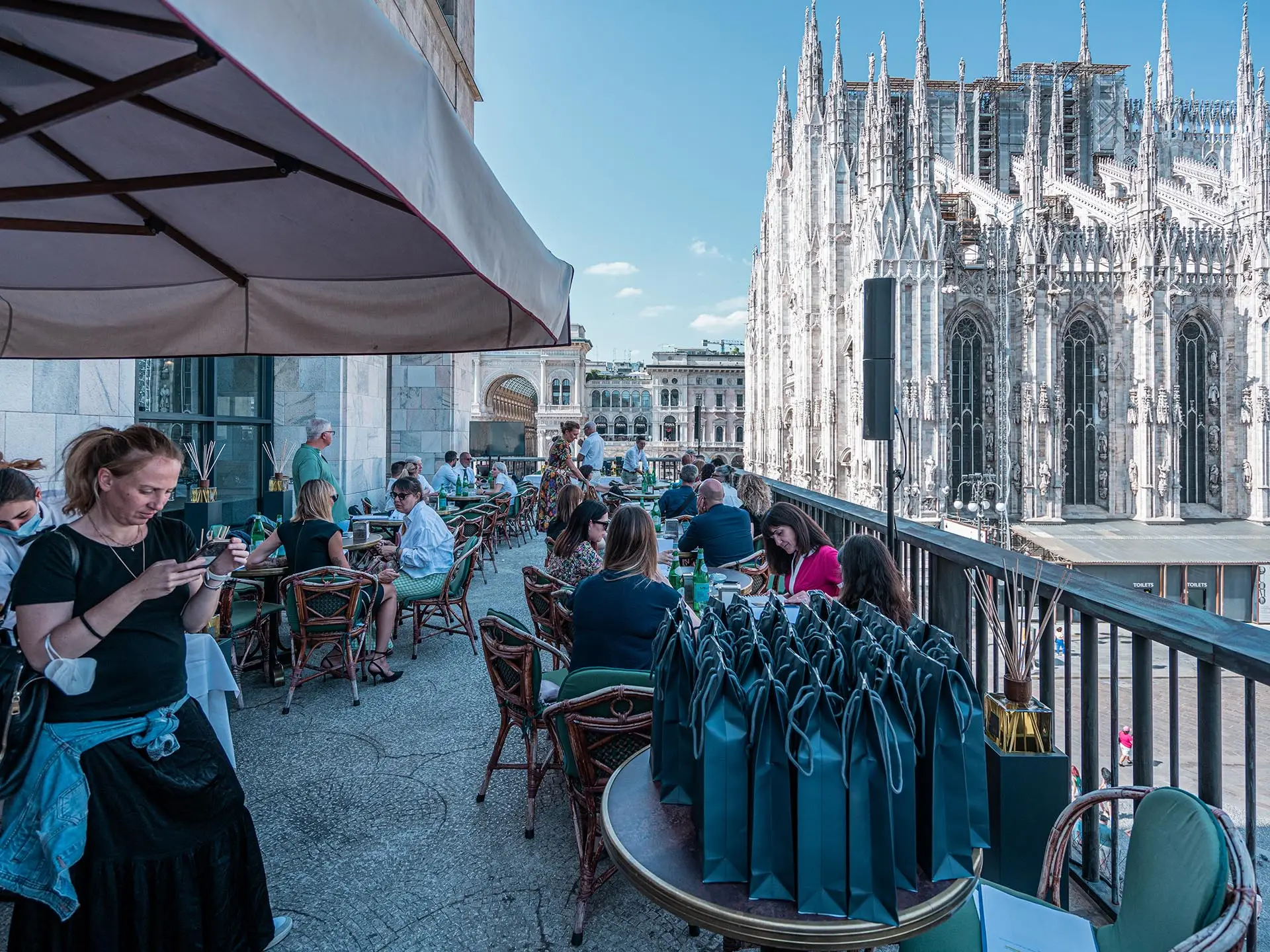 Evento di presentazione della fragranza Duomo Milano di Euthalia Fragrances presso Terrazza Giacomo Arengario, con vista panoramica sul Duomo di Milano. Invitati che partecipano all'evento, seduti ai tavoli all'aperto, con borse regalo su un tavolo in primo piano. Atmosfera elegante e rilassata, perfetta per scoprire la nuova fragranza ispirata alla maestosità del Duomo.