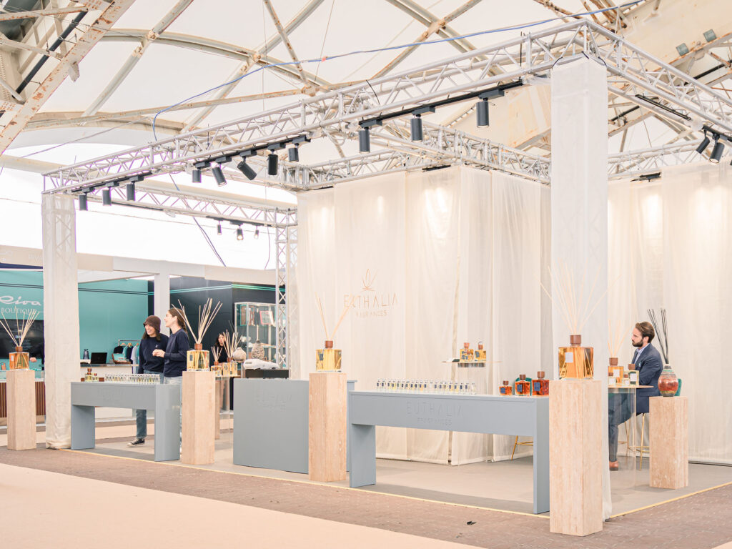 Stand Euthalia Fragrances al Salone Nautico di Genova 2024, con esposizione elegante di diffusori di profumo e fragranze di lusso. Lo stand, caratterizzato da un design moderno e raffinato, include profumi esclusivi e diffusori in vetro decorativo, con professionisti del settore intenti a interagire con i visitatori. Atmosfera luminosa e spaziosa, che riflette l'alta qualità dei prodotti esposti. 64th Genoa Boat Show.