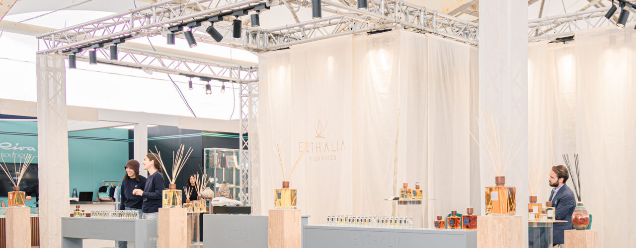 Stand Euthalia Fragrances al Salone Nautico di Genova 2024, con esposizione elegante di diffusori di profumo e fragranze di lusso. Lo stand, caratterizzato da un design moderno e raffinato, include profumi esclusivi e diffusori in vetro decorativo, con professionisti del settore intenti a interagire con i visitatori. Atmosfera luminosa e spaziosa, che riflette l'alta qualità dei prodotti esposti. 64th Genoa Boat Show.