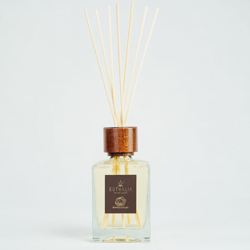 Fragranza Bianco d'Alba. Diffusore a bastoncini Euthalia Fragrances, con flacone in vetro trasparente. Il tappo in legno e i bastoncini completano il design elegante, ideale per profumare ambienti con una fragranza raffinata e di lunga durata.