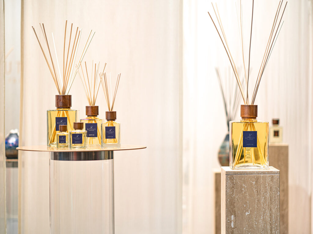 Protagonista della scena, il diffusore a bastoncini di Euthalia Fragrances si distingue per il suo design elegante e sofisticato. Esposti su piedistalli e superfici raffinate, i flaconi di diverse dimensioni, con tappo in legno e bastoncini in rattan, diffondono un'aura di lusso e armonia nell'ambiente. Lo sfondo neutro e le luci soffuse esaltano la bellezza delle fragranze, trasformando lo stand in un'esperienza sensoriale unica.