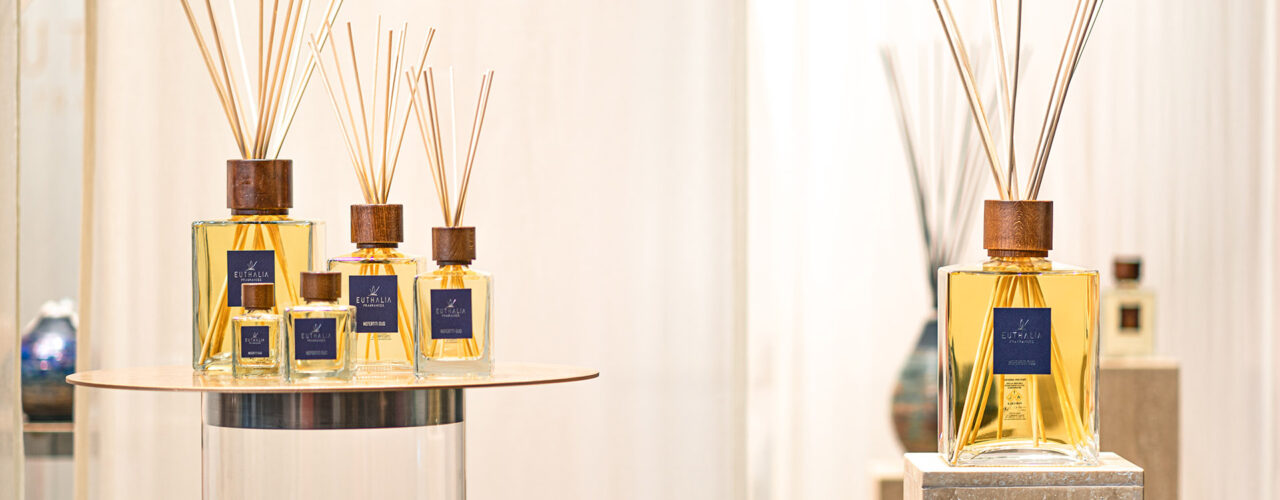 Protagonista della scena, il diffusore a bastoncini di Euthalia Fragrances si distingue per il suo design elegante e sofisticato. Esposti su piedistalli e superfici raffinate, i flaconi di diverse dimensioni, con tappo in legno e bastoncini in rattan, diffondono un'aura di lusso e armonia nell'ambiente. Lo sfondo neutro e le luci soffuse esaltano la bellezza delle fragranze, trasformando lo stand in un'esperienza sensoriale unica.